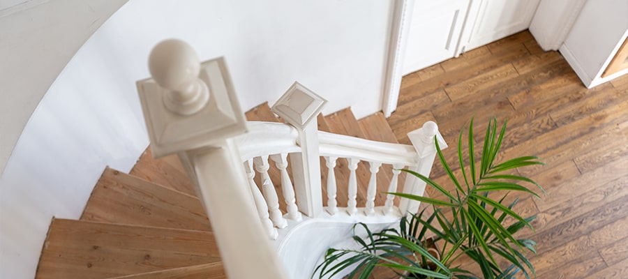 Eine Draufsicht auf eine Holztreppe mit weißen Geländerstäben und einem Handlauf, neben einer grünen Topfpflanze, die hinunter zu einem Holzboden mit Fischgrätenmuster führt.