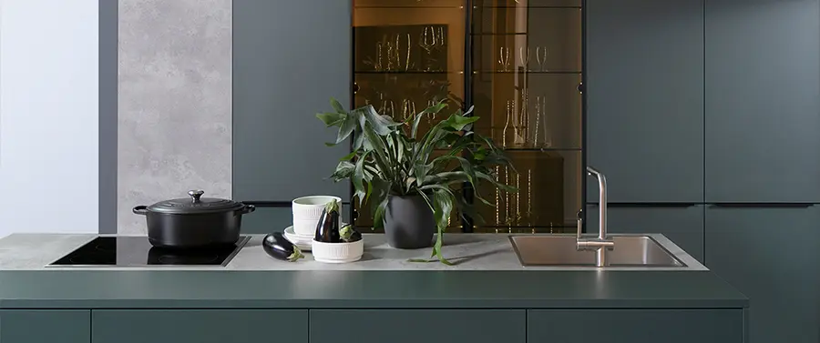 Eine moderne Design-Küchentheke mit einem schwarzen Topf, einer Tasse mit Utensilien und einer grünen Topfpflanze, daneben eine Edelstahlspüle, mit dunkelgrünen Schränken und einer Glasfront