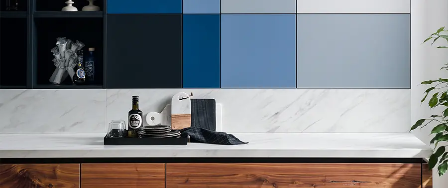 Eine moderne Küchenarbeitsplatte von Küchenstile mit Marmoroberfläche und Holzschranksockel sowie einer geometrisch gemusterten Rückwand aus blauen und grauen Fliesen. Zum Zubehör gehören Teller, eine Flasche und ein Glas