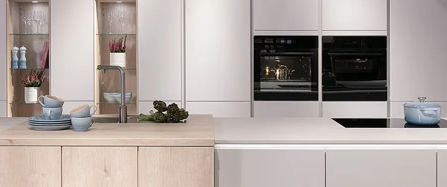 Ein modernes Kücheninterieur mit eleganten, weißen Schränken, einer Holzarbeitsplatte mit Essgeschirr, Einbaugeräten und Küchenausstattung in den Regalen.