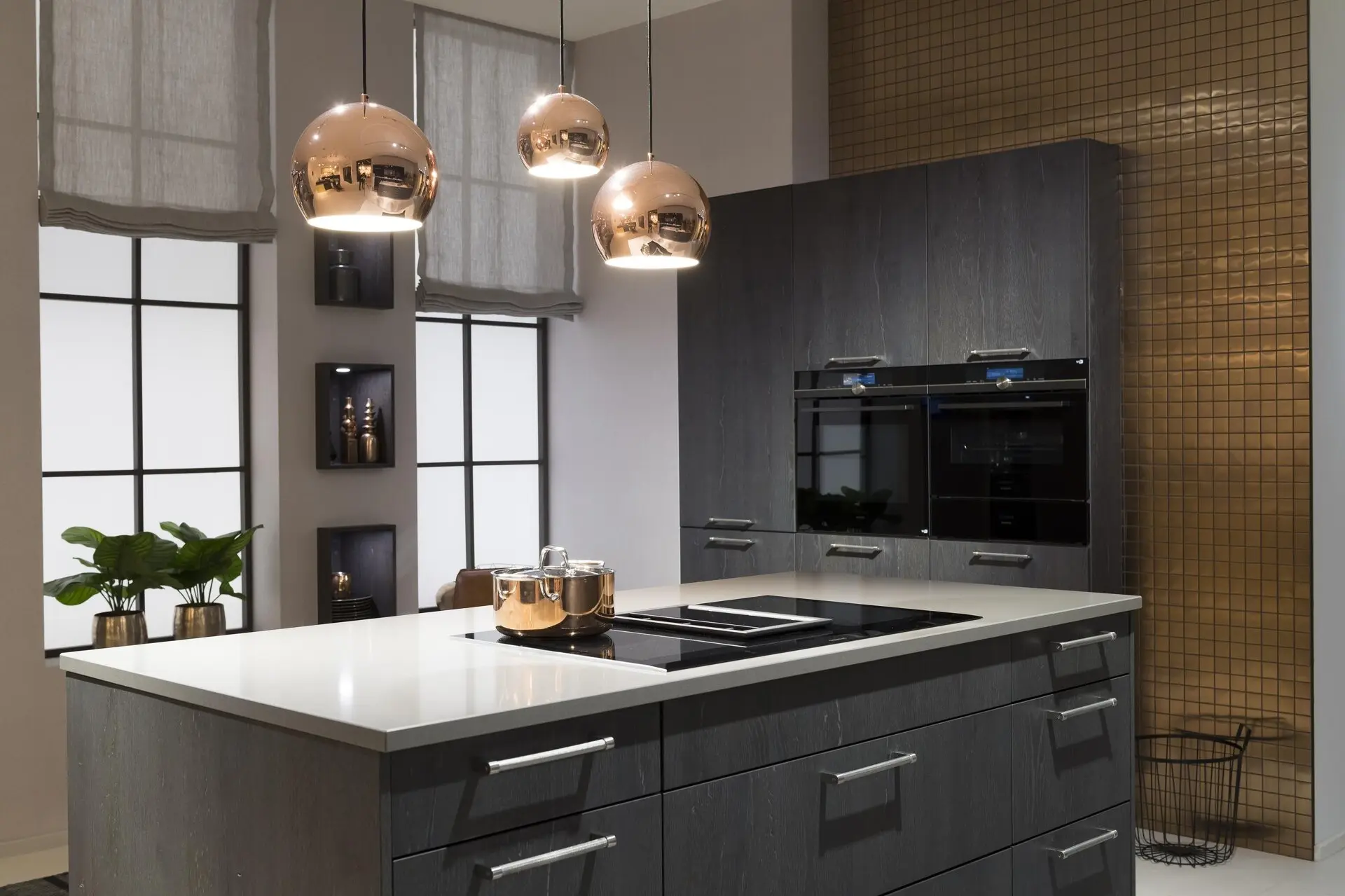 Modernes Kücheninterieur mit dunklen Häcker-Schränken, Küchengeräten aus Edelstahl und kupferfarbenen Pendelleuchten über einer weißen Kücheninsel.