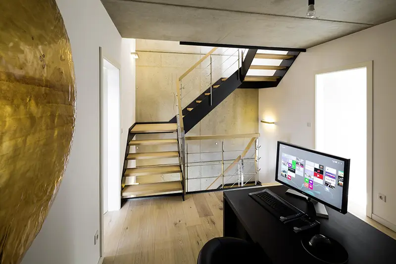 Ein modernes Heimbüro mit Computereinrichtung, HPL-Treppen, das zu einem oberen Stockwerk auf der linken Seite führt, und einer großen, strukturierten goldenen Dekoration an der Wand.