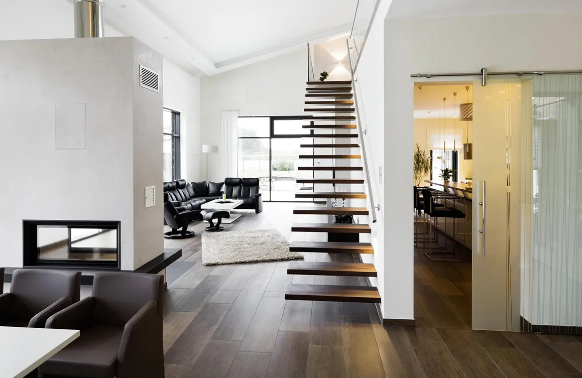 Ein modernes Wohnzimmer mit dunklen Holzböden, einer schwebenden Kragarmtreppe und einem offenen Grundriss, der zu einem Küchenbereich führt.