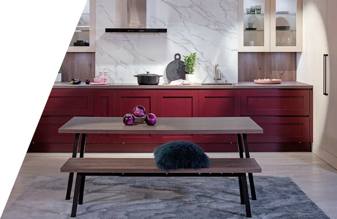 Moderne Küche mit Marmorrückwand, dunkelroten Schränken und einer Mittelinsel, begleitet von einem Holztisch mit Bank und Dekorationsgegenständen über einem grau gemusterten Teppich mit Küchenpflege