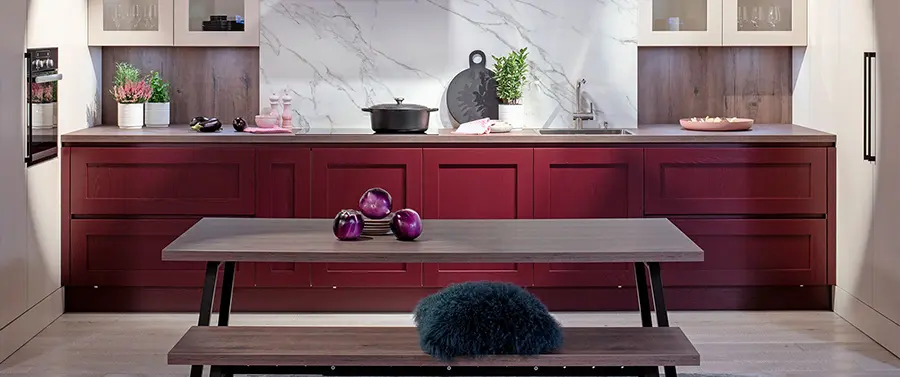 Moderne Küche mit roten Holzschränken, Marmorrückwand und einem Holztisch mit Bänken, akzentuiert durch lila Dekoobjekte und ein blaues Fellkissen von Küchenstudio Melle.