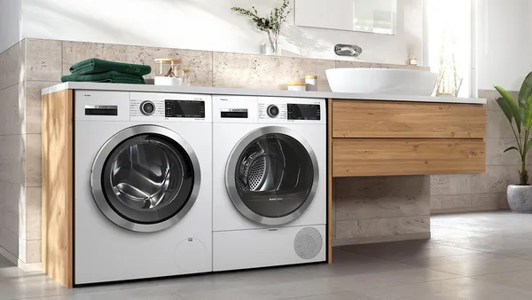 Ein moderner Waschraum mit einer weißen Bosch-Frontlader-Waschmaschine und einem Trockner, die nebeneinander unter einer Holzarbeitsplatte platziert sind, neben einem Waschbecken im Badezimmer, auf dem ein grünes Handtuch liegt.