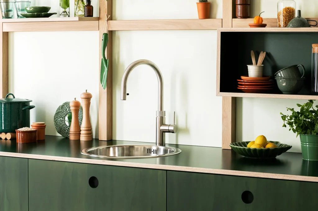 Moderne Küchentheke mit Edelstahlspüle und Quooker, grünen Schränken und Holzregalen mit verschiedenen Küchenutensilien und Pflanzen.