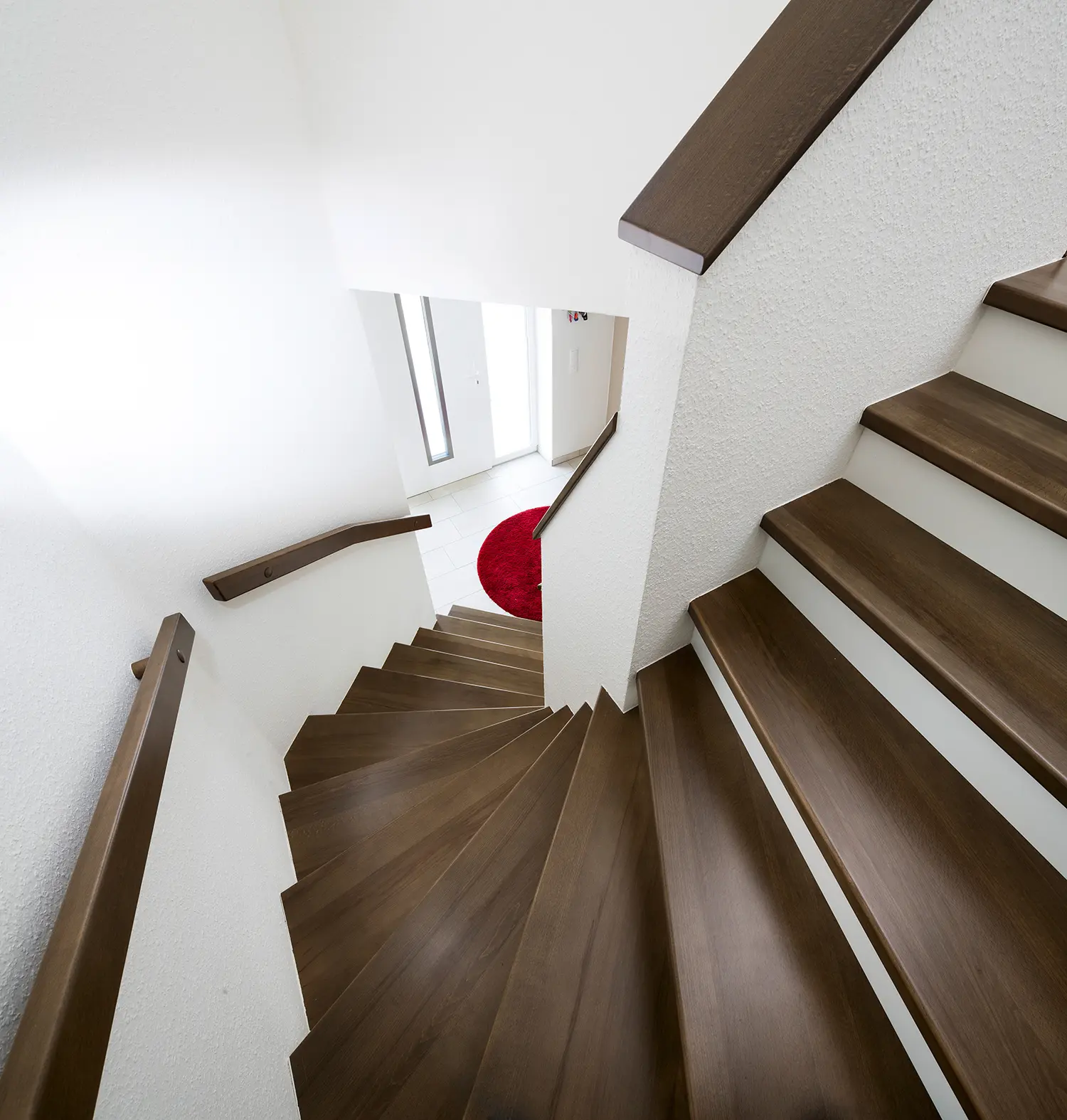 Innenansicht einer modernen Treppe mit Stufen aus dunklem Holz, die zu einem Boden mit einem roten runden Teppich führt, mit weißen Wänden und Handläufen auf Beton.