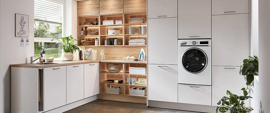 Ein modernes Kücheninterieur mit weißen Schränken auf der rechten Seite und einer eingebauten Waschmaschine, offenen Holzregalen auf der linken Seite und einer Arbeitsplatte mit verschiedenen Küchenutensilien und dekorativen Pflanzen für eine harmonische Atmosphäre