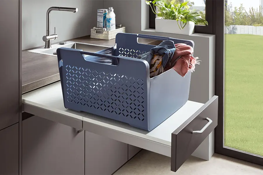 Ein mit Kleidung gefüllter Wäschekorb steht auf einer offenen Schublade neben einem Spülbecken im Hauswirtschaftsraum, auf der Arbeitsplatte ist eine Flasche Waschmittel sichtbar.