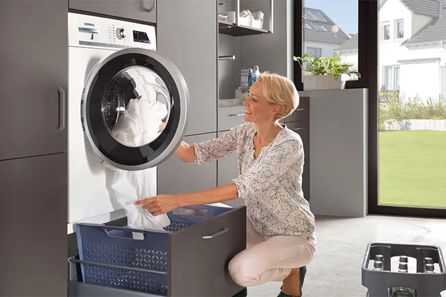 Eine Frau zieht Wäsche aus einer Frontlader-Waschmaschine, die im Hauswirtschaftsraum auf einem Podest mit ausziehbarer Schublade steht.