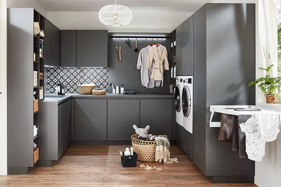 Eine kompakte, moderne Waschküche mit dunkelgrauen Schränken, einer Waschmaschine und einem Trockner sowie einer gemusterten Rückwand und Holzboden. In den Hauswirtschaftsräumen hängt Kleidung zum Trocknen