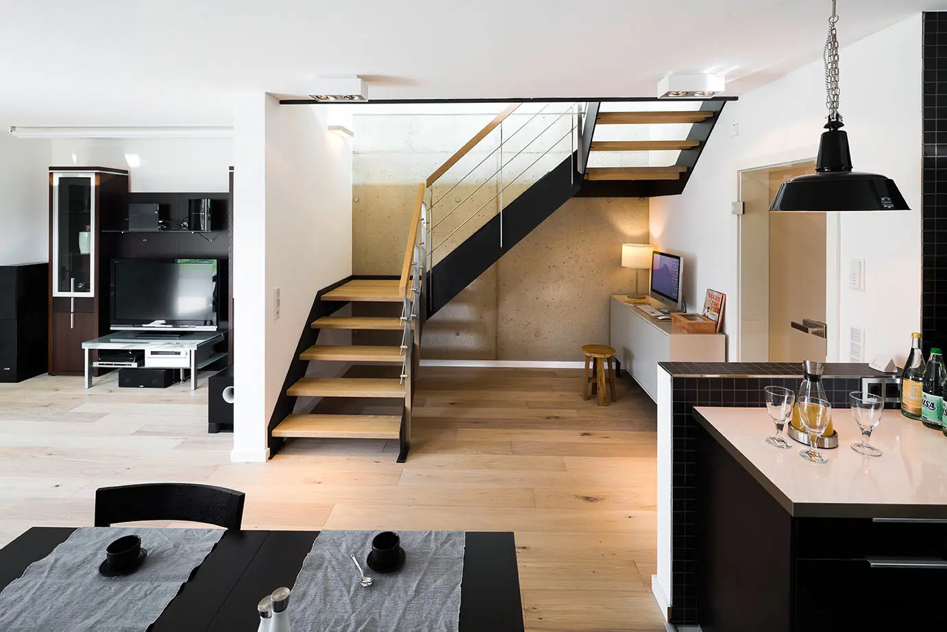 Ein modernes Kücheninterieur mit einer Treppe, die zu einer oberen Ebene führt, mit einem schwarzen Esstisch, einer Kücheninsel mit Barhockern und einem offenen Grundriss. Die Treppe ist hochwertig verarbeitet