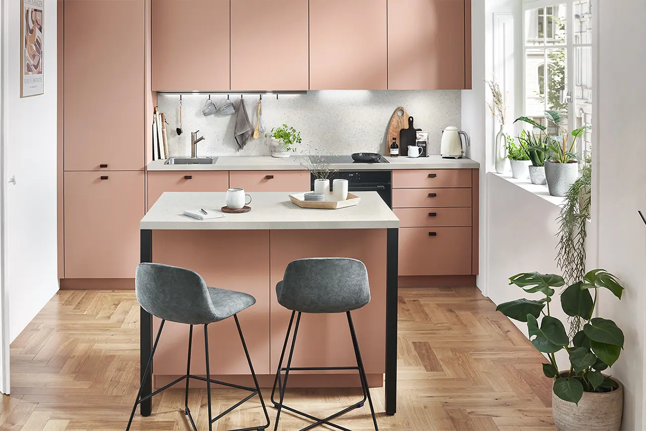 Eine moderne, individuelle Küche mit rosafarbenen Schränken, einer zentralen Insel mit zwei grauen Stühlen und Zimmerpflanzen am Fenster