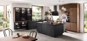 Ein Mann steht in einer modernen Küche mit dunklen Schränken und nippt aus einer Tasse neben einem offenen Kühlschrank, mit einer Kücheninsel im Vordergrund und einem gut beleuchteten Essbereich mit klassischer Küche