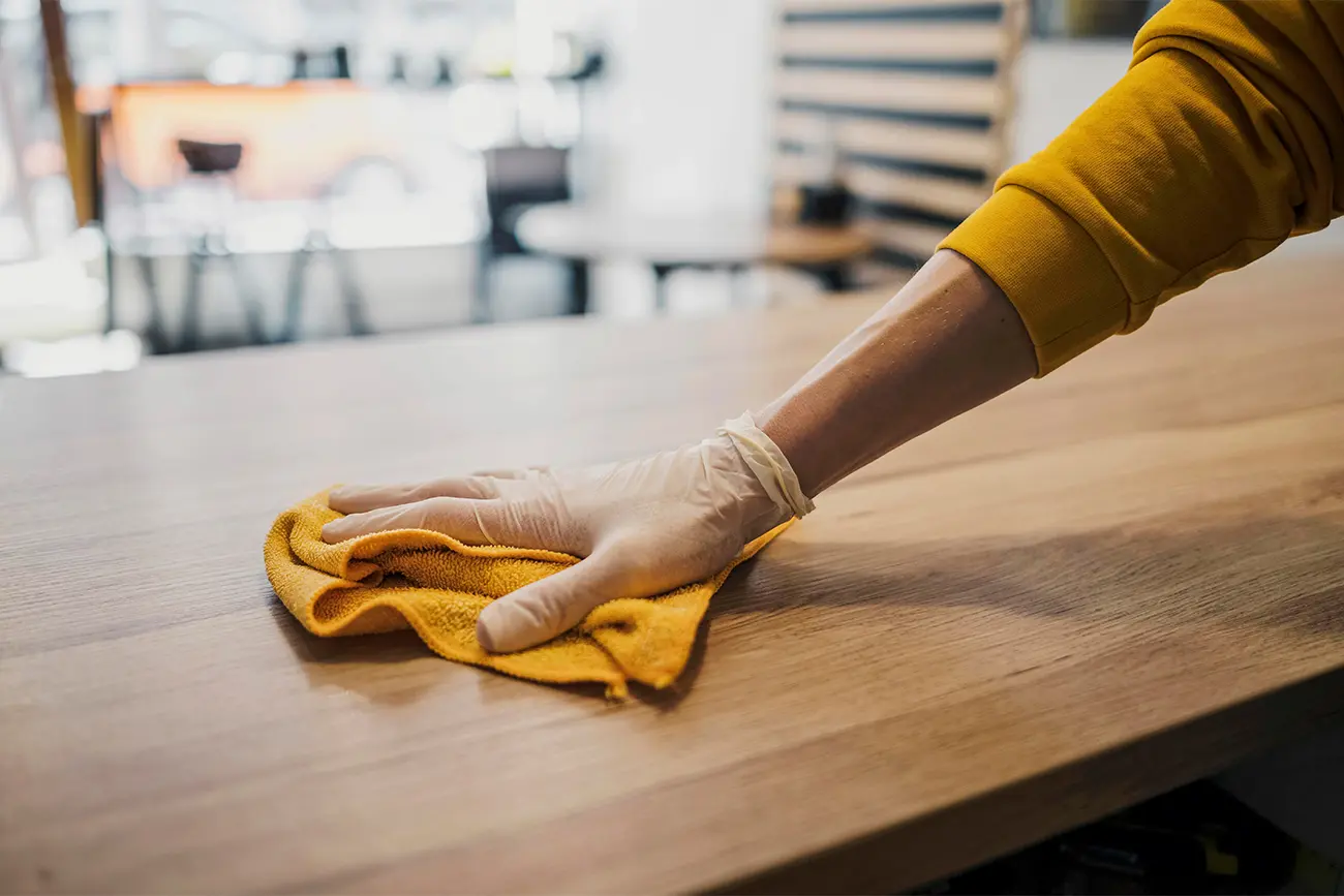 Eine Person im gelben Hemd und mit weißen Handschuhen wischt zur Küchenpflege einen Holztisch mit einem gelben Tuch ab.