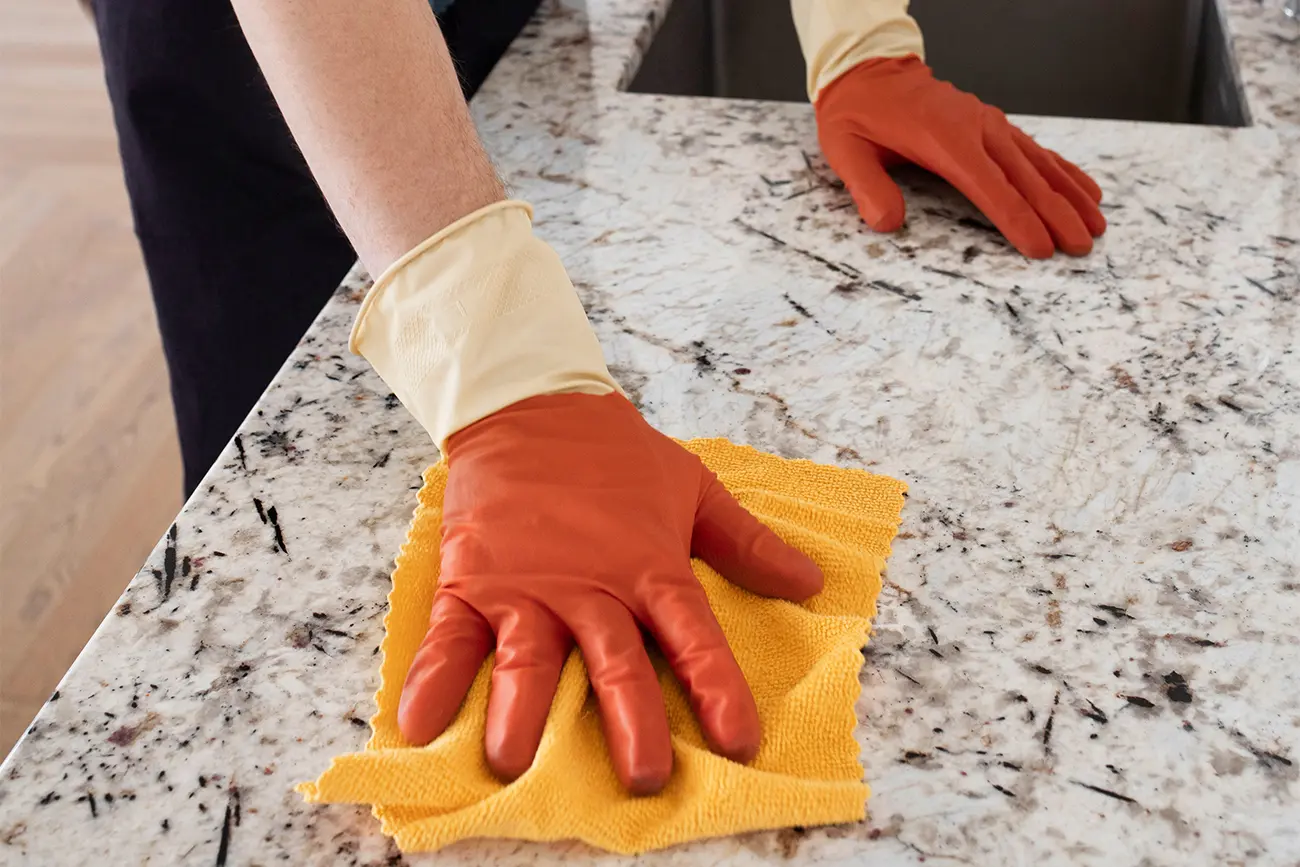 Eine Person mit orangefarbenen Gummihandschuhen wischt zur Küchenpflege eine Marmorarbeitsplatte mit einem gelben Tuch ab.