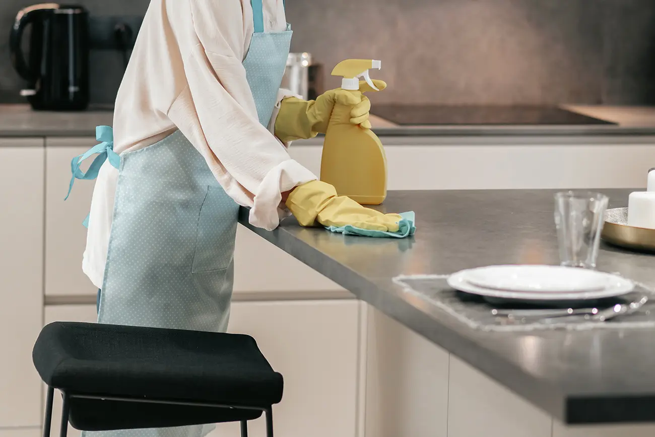 Eine Person mit gelben Handschuhen und einer hellblauen Schürze mit Schleife führt Küchenpflege durch, indem sie eine Küchenarbeitsplatte mit einem Tuch reinigt und in der anderen Hand eine Sprühflasche hält. Dort
