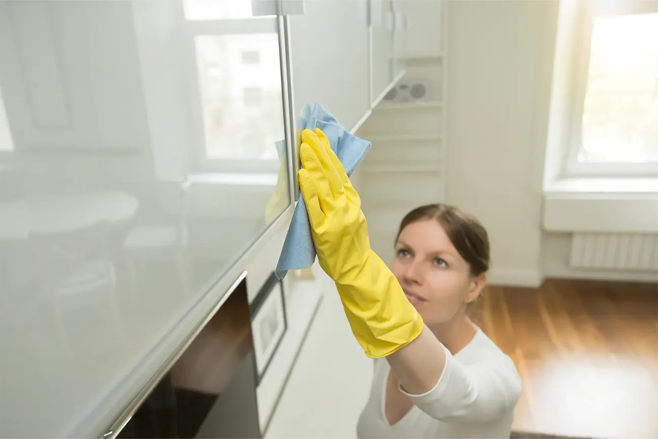 Eine Person mit gelben Handschuhen reinigt die Vorderseite eines weißen Küchenschranks mit einem blauen Tuch und übt dabei engagierte Küchenpflege aus.