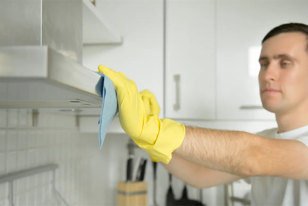 Eine Person mit gelben Handschuhen führt Küchenpflege durch, indem sie eine Dunstabzugshaube mit einem blauen Tuch reinigt.