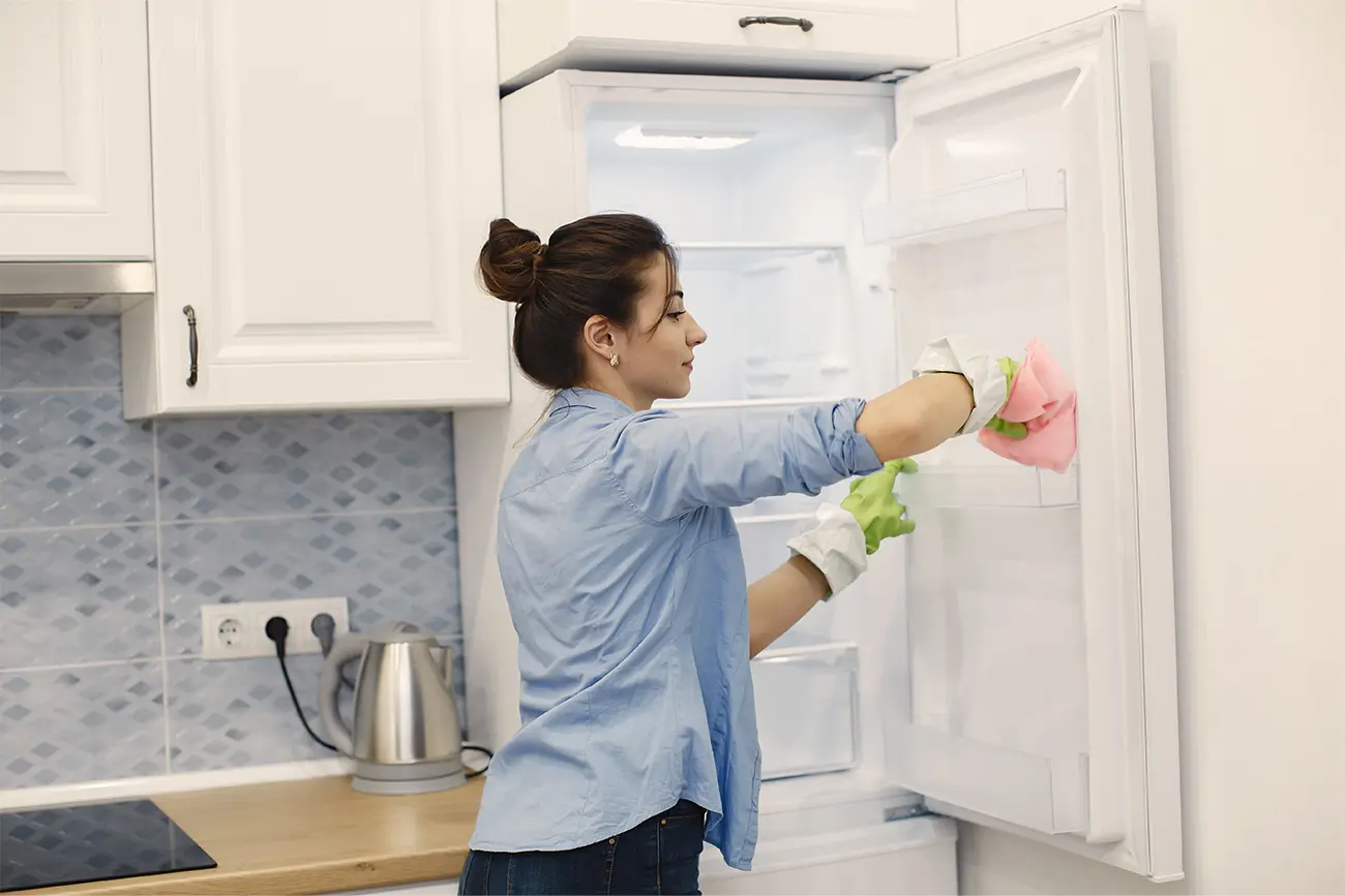 Eine Frau in einem blauen Hemd führt Küchenpflege durch, indem sie das Innere eines offenen Kühlschranks mit einem Tuch reinigt.