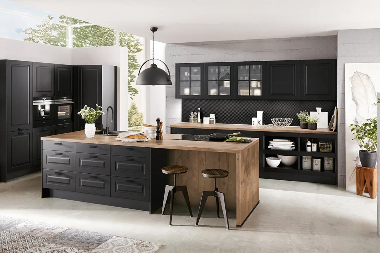 Eine moderne Landhausküche mit schwarzen Schränken, Küchengeräten aus Edelstahl, einer Holzinsel mit Sitzgelegenheiten und weißen Arbeitsplatten, die den Raum mit natürlichem Licht erhellen.
