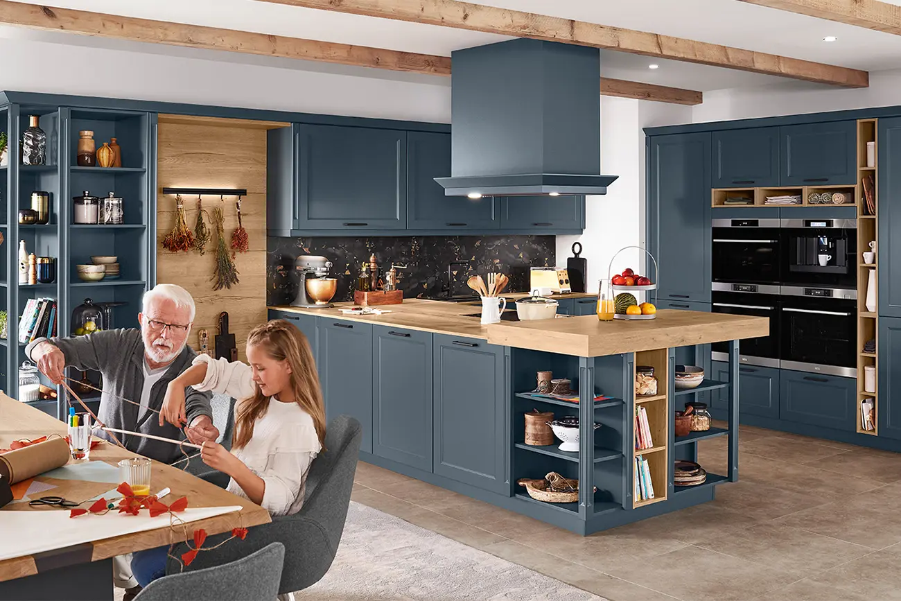 Ein älterer Mann und ein junges Mädchen basteln auf einer Kücheninsel in einer Landhausküche mit blauen Schränken und Geräten aus Edelstahl.