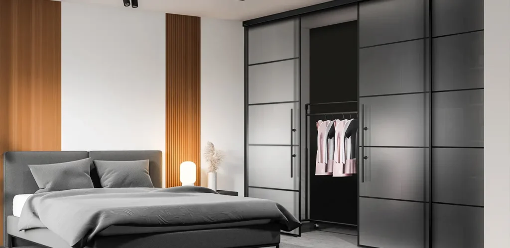 Ein modernes Schlafzimmer mit einem großen Bett mit grauer Bettwäsche, einem Kopfteil aus Holz, einem Nachttisch mit Lampe und einem schwarzen Kleiderschrank mit geöffneten Glastüren, in dem mehrere hängende Kleidungsstücke ausgestellt sind.