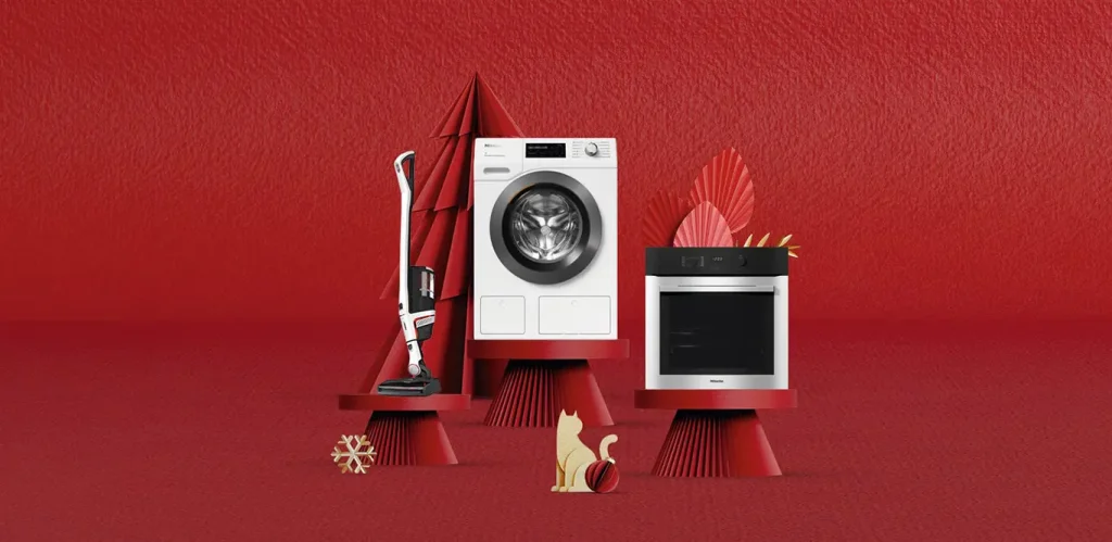 Ein Miele-Staubsauger, eine Waschmaschine und ein Mikrowellenherd auf roten Sockeln vor einem roten Hintergrund mit dekorativen geometrischen Formen, einer Schneeflocke und einer Eichhörnchenfigur.