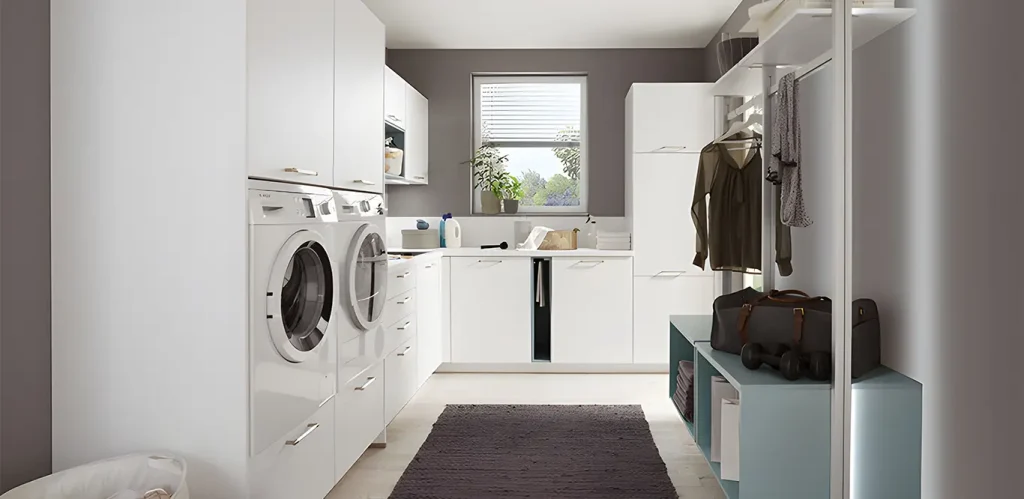 Ein moderner Waschraum mit weißen Schränken, Waschmaschine, Trockner und einem Waschbecken unter einem Fenster mit geschlossener Jalousie, dazu ordentlich aufgehängte Kleidung und ein paar Wäscheartikel
