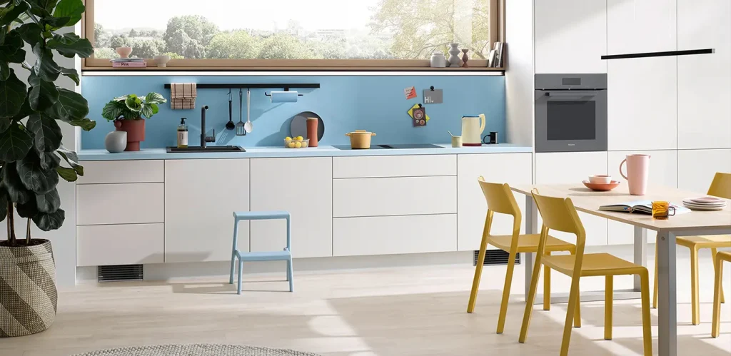 Eine moderne Küche mit weißen Schränken, Esszimmermöbeln aus hellem Holz und hellem Naber-Küchenzubehör auf der hellblauen Rückwand, akzentuiert mit einer Vielzahl von Pflanzen und Akzenten