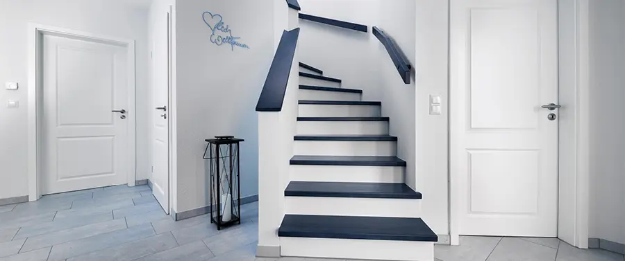 Eine moderne Treppe mit dunklen Stufen vor weißen Setzstufen führt zur oberen Ebene eines sauberen und minimalistischen Innenraums mit hellgrauen Wänden und Fliesenboden.