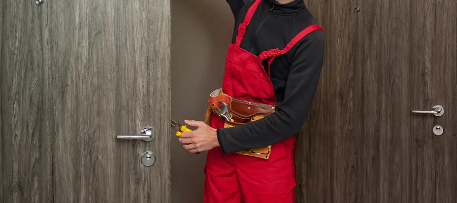 Eine Person in einem roten Overall und einem schwarzen Kapuzenpullover mit Werkzeuggürtel öffnet eine graue Holztür, die ihr Gesicht teilweise vor der Kamera verdeckt und in einen der Innenräume des Gebäudes führt