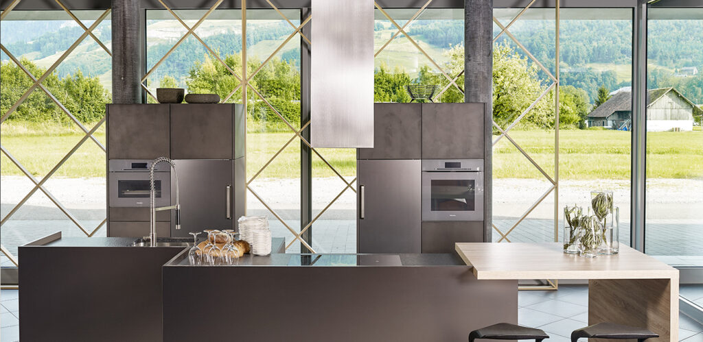 Modernes Kücheninterieur mit klaren Linien und Einbaugeräten, mit Blick auf eine ruhige Außenlandschaft durch ein großes geometrisches Fenster. Diese „Individuelle Küche“ verbindet Stil und Funktionalität nahtlos.