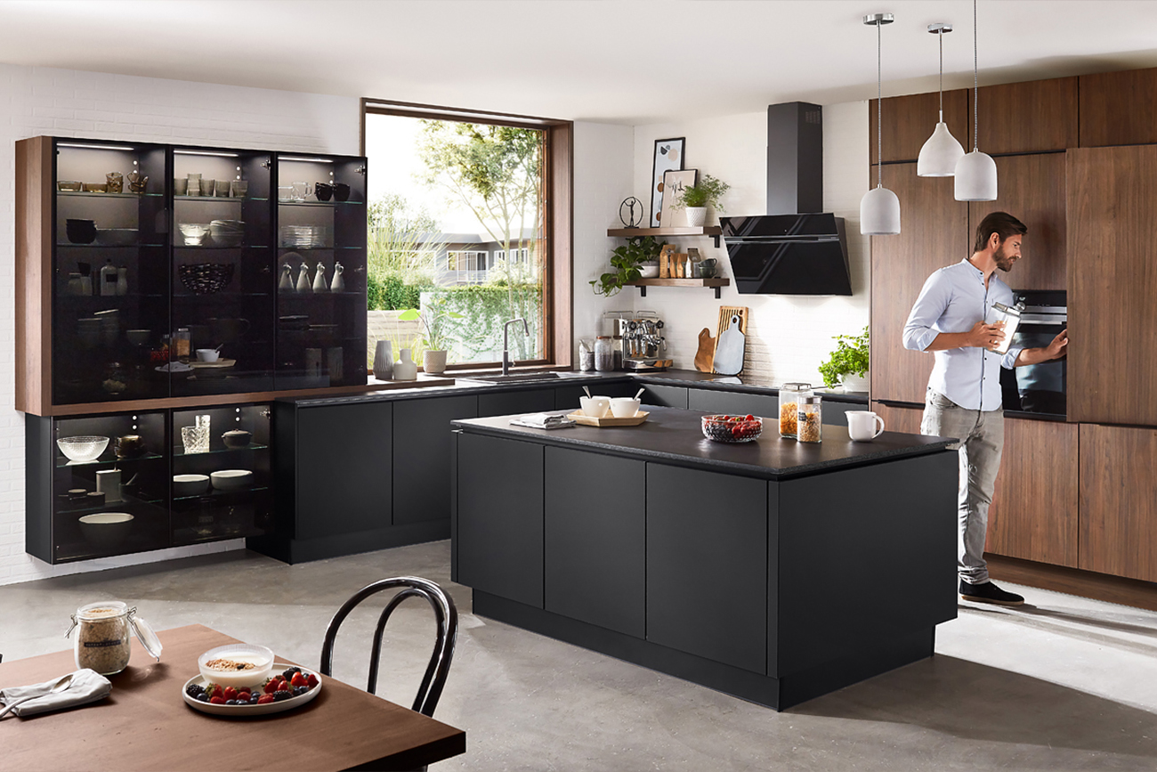 Ein Mann steht in einer klassischen Küche mit dunklen Schränken und interagiert mit einem Kühlschrank, während die Kücheninsel und die Arbeitsflächen verschiedene Kochutensilien und Zutaten präsentieren.