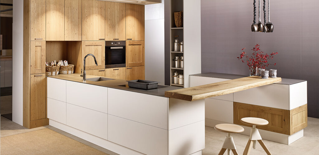 Modernes Kücheninterieur mit weißen Schränken, Holzakzenten und integrierten Geräten von Natural Living Küchen, ergänzt durch minimalistische Barhocker und dekorative rote Zweige auf der Arbeitsplatte.